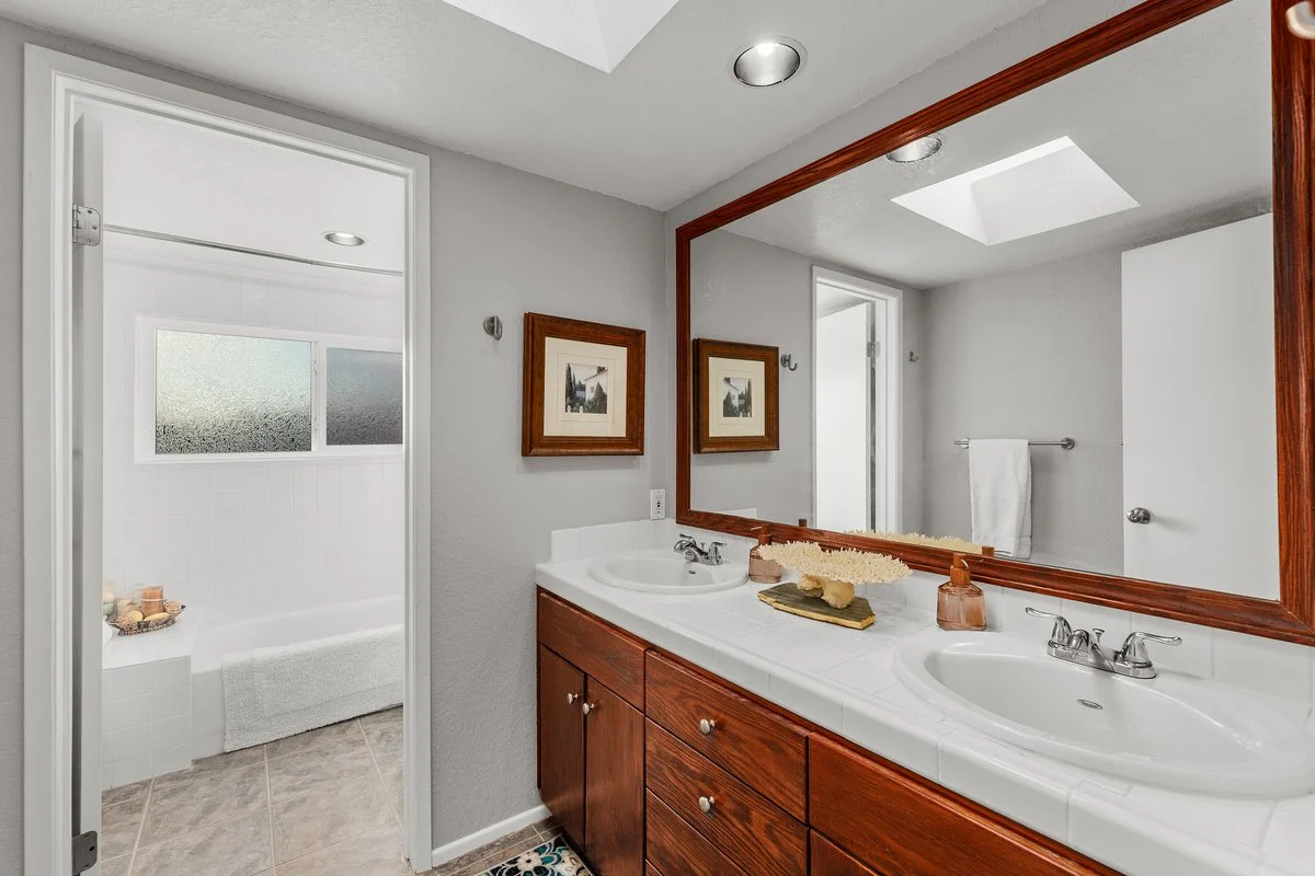 A bathroom with a double sink vanity featuring a large mirror with a wooden frame, framed pictures on the wall, a towel bar with a white towel, and a walk-in shower with a frosted window and a bathtub, small decorative items on the counters.