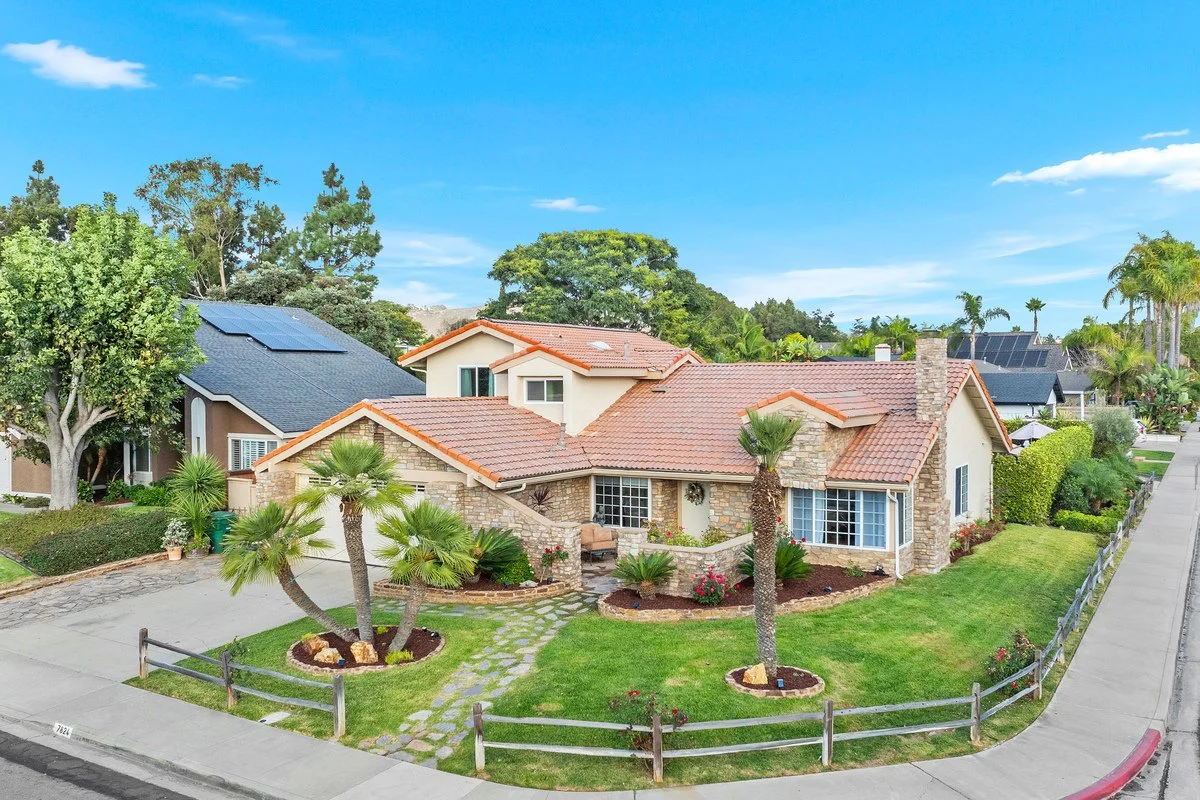 A suburban house with a red tile roof, surrounded by a well-manicured lawn with palm trees and shrubbery, and neighboring houses, under a blue sky.