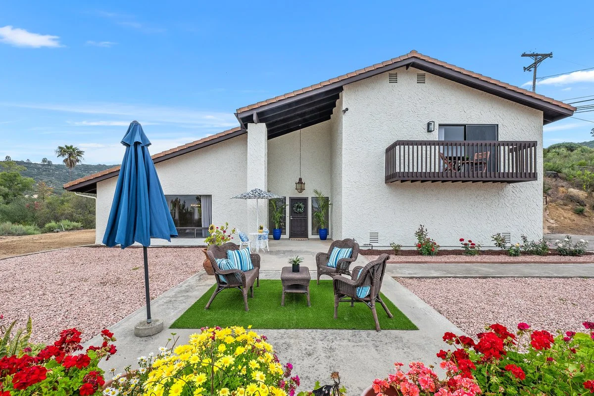 A two-story house with a white stucco exterior, brown roof, and a small balcony on the second floor. In front, there is a patio area with outdoor seating, a large blue umbrella, a green artificial grass area, and colorful flowers.