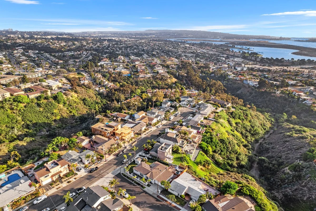 Aerial view of a suburban neighborhood nestled on a hillside, with houses, streets, trees, and a body of water in the background under a partly cloudy sky.