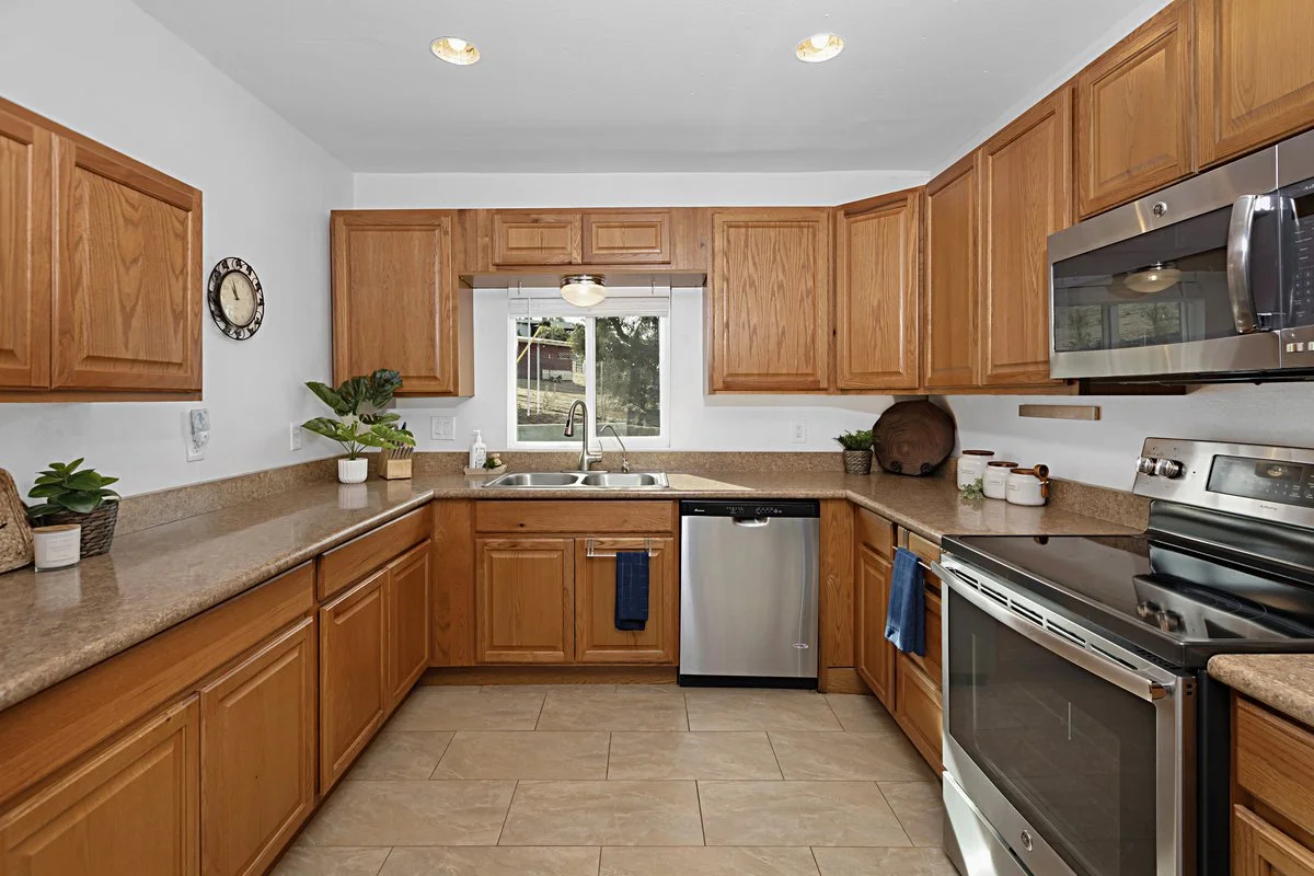 Kitchen with wooden cabinets, beige countertops, stainless steel appliances including a microwave, oven, and dishwasher, and a window above the sink. There are potted plants and a clock on the walls.