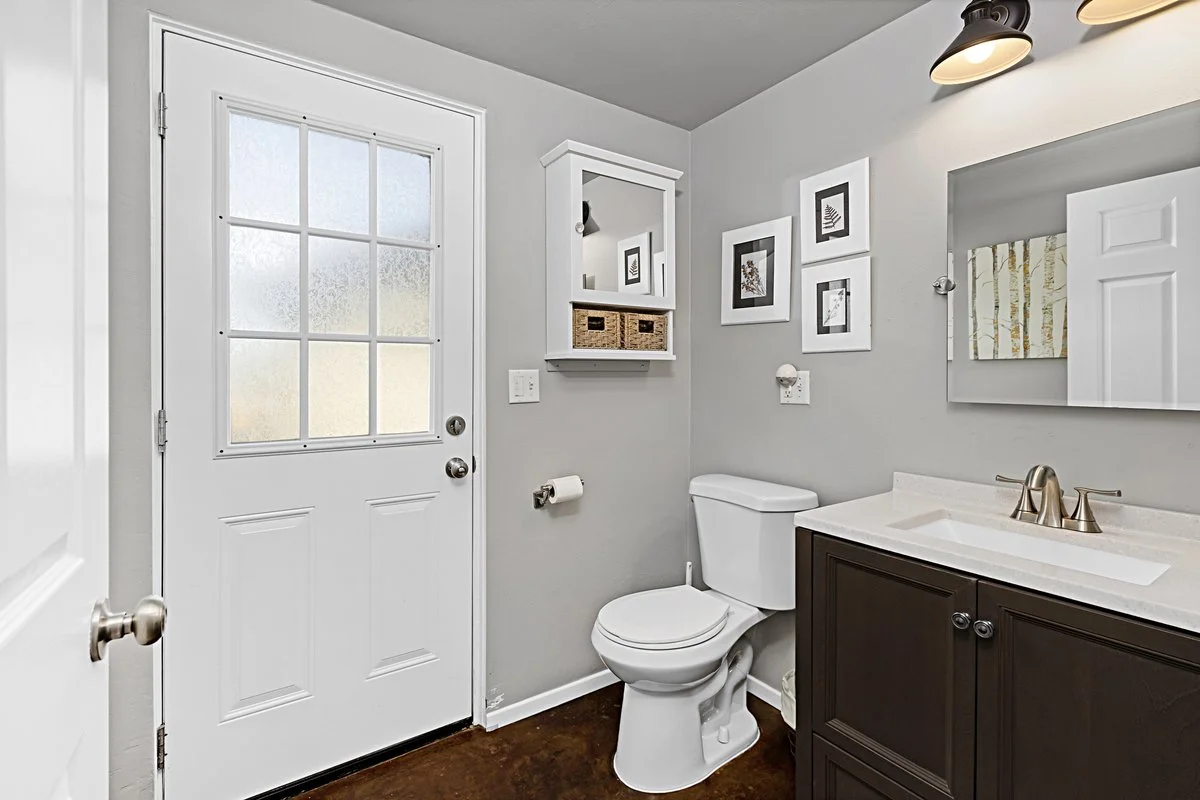 Bathroom with a white door, a toilet, a dark brown vanity with a white countertop, and a mirror, with framed artwork and a small wall cabinet.