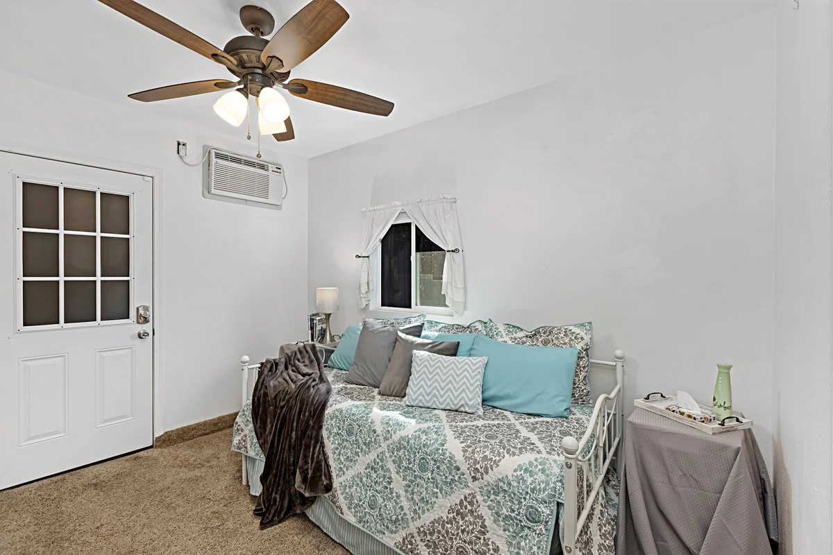 A bedroom with a white bedspread, multiple pillows, a ceiling fan with lights, a small window with white curtains, and a table covered with a purple cloth holding a green vase and other items.