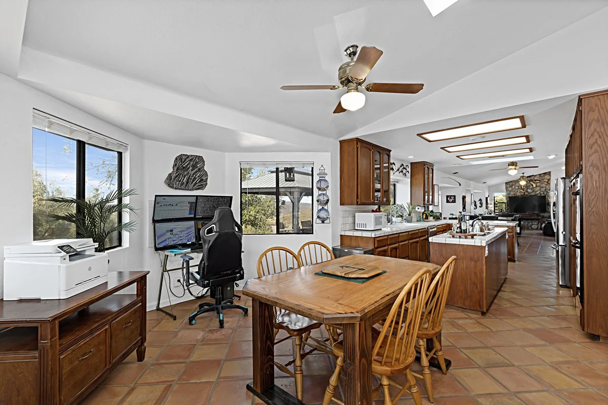 Open-concept kitchen and dining area with wood cabinets, tiled floor, ceiling fans, and a desk with dual monitors near a window.