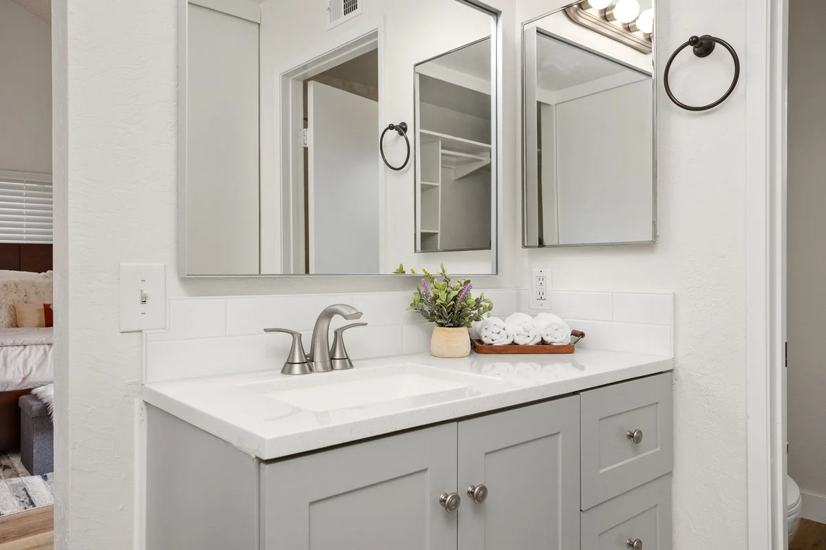 Bathroom vanity with a white countertop, two cabinet doors, and two drawers, a silver faucet, a potted plant, and a tray with rolled white towels. Two mirrors, one with a reflection of the shower area, and two towel rings are mounted on the wall.