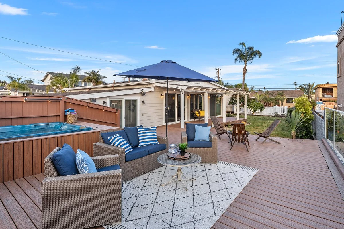 Backyard deck with outdoor furniture, hot tub, sun umbrella, seating area with striped cushions, and palm trees under a clear blue sky.
