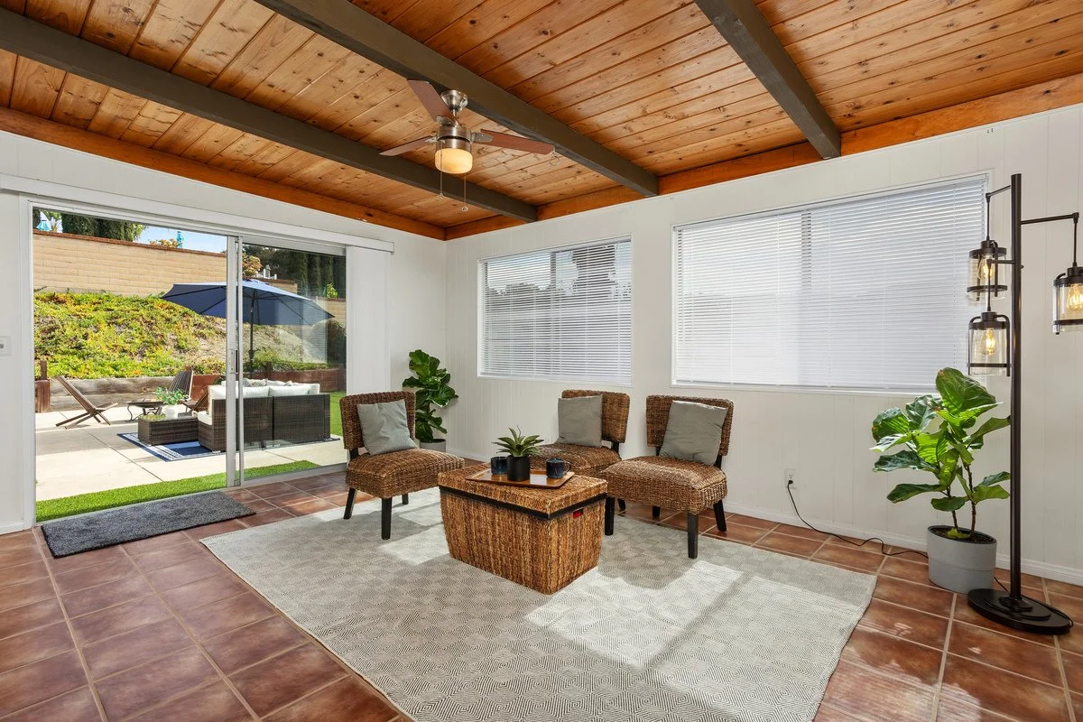 Living room with wooden ceiling, tile floor, wicker chairs, glass sliding door leading outside to patio, potted plants, and large windows with blinds.