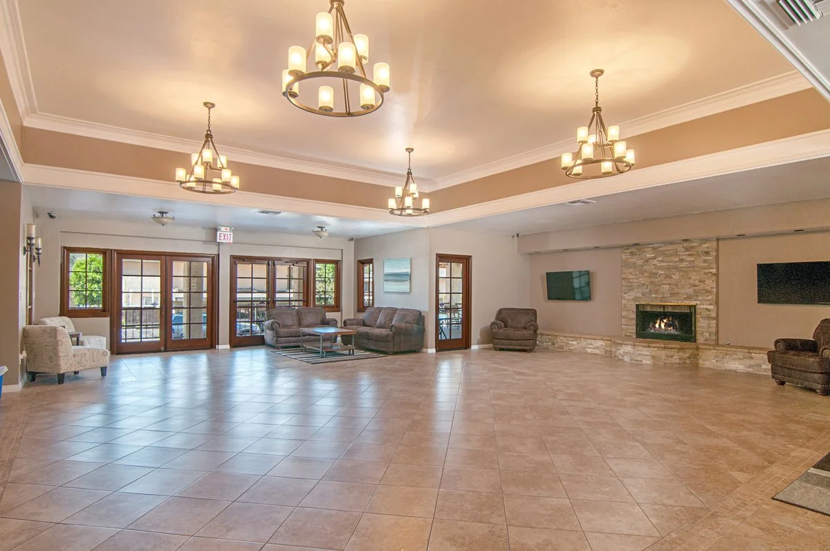 Spacious lounge area with tiled floor, multiple chandeliers, a fireplace, wall-mounted TV, and several armchairs and sofas near large windows and glass doors.