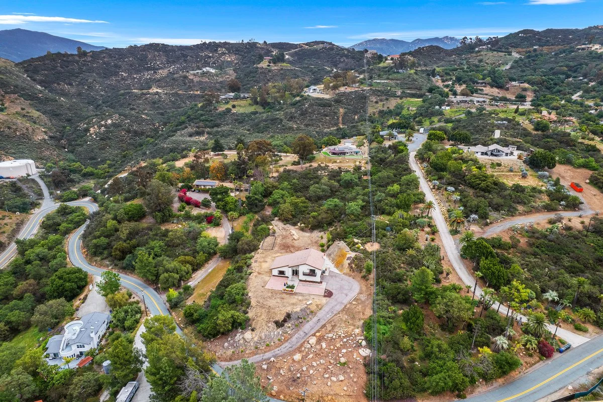 Aerial view of a mountainous residential area with winding roads, houses, and lush greenery under a blue sky.