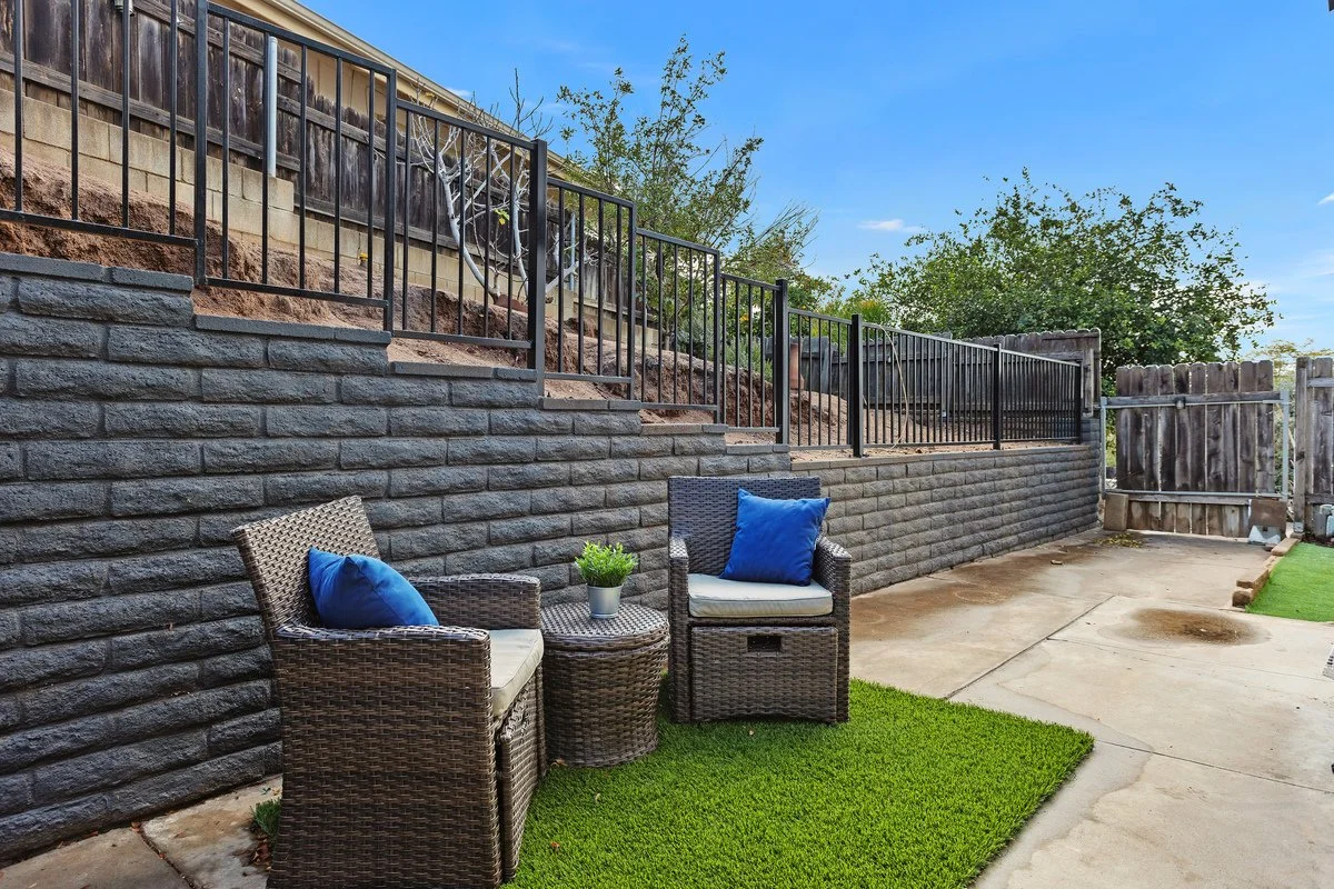 Backyard patio with two wicker chairs with blue pillows, a small wicker table with a potted plant, green artificial grass, a concrete patio, a stone retaining wall, metal railing, and a wooden fence in the background under a clear blue sky.