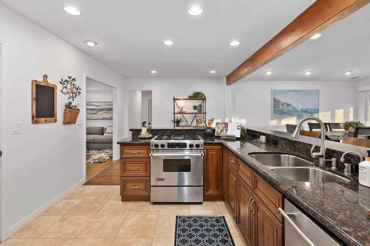 Modern kitchen with stainless steel oven and microwave, brown wooden cabinets, granite countertops, and a double sink. The room has recessed lighting, a decorative wall with hanging plants and chalkboard, and a view into a living room and dining area