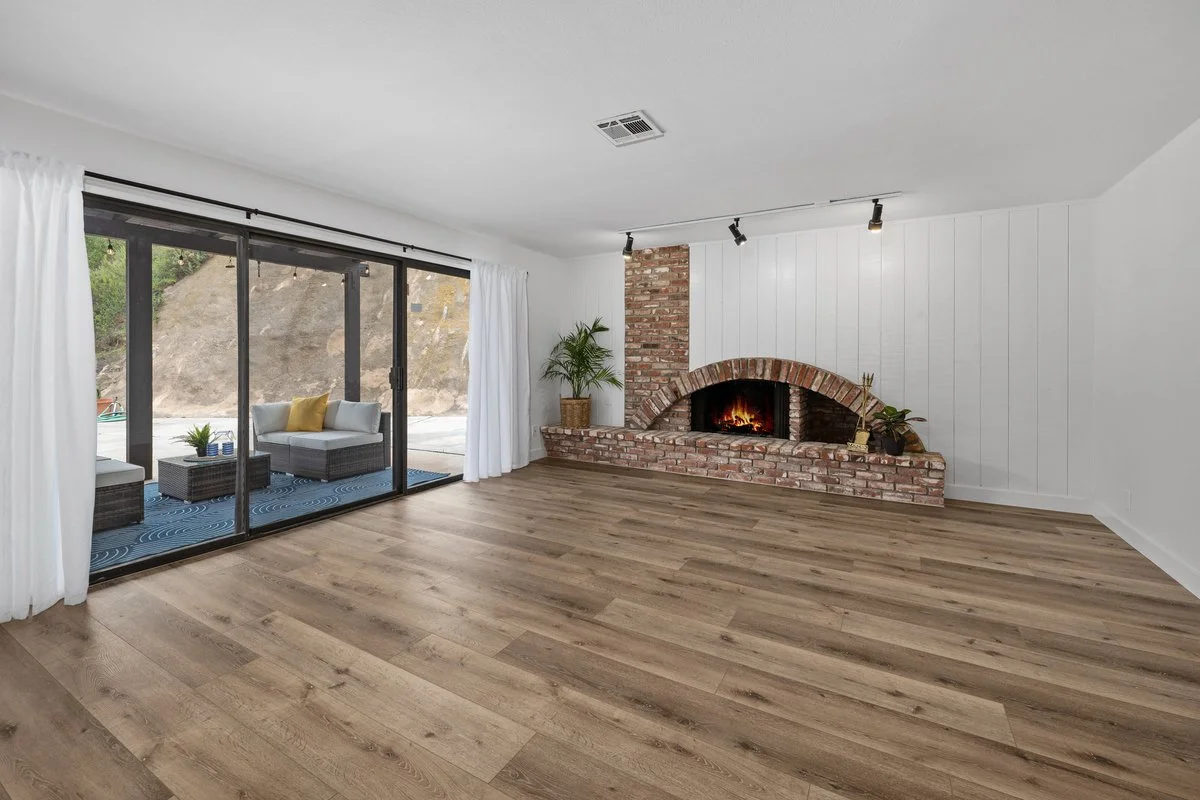Empty living room with hardwood floor, brick fireplace, sliding glass door with white curtains, and outdoor seating area with two chairs and a table.