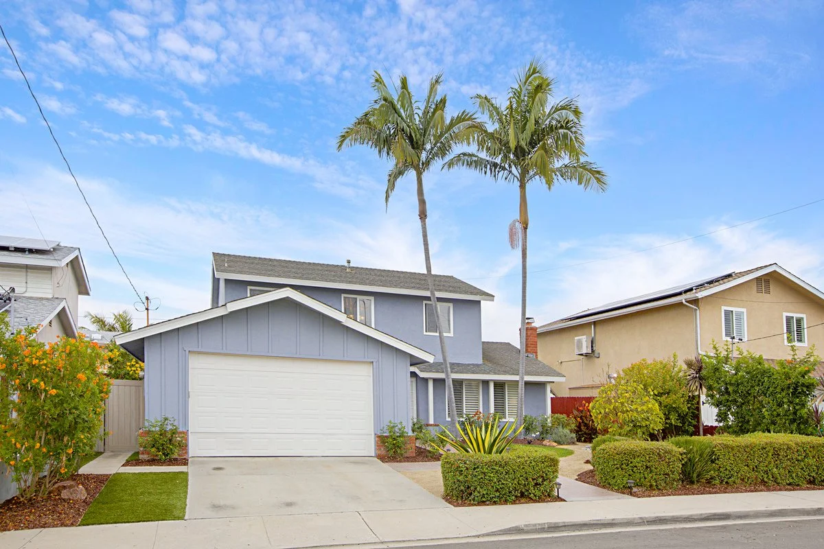 A two-story light blue house with a gray roof, white garage door, two tall palm trees in the front yard, and well-maintained bushes and plants, located on a sunny day with a partly cloudy sky.