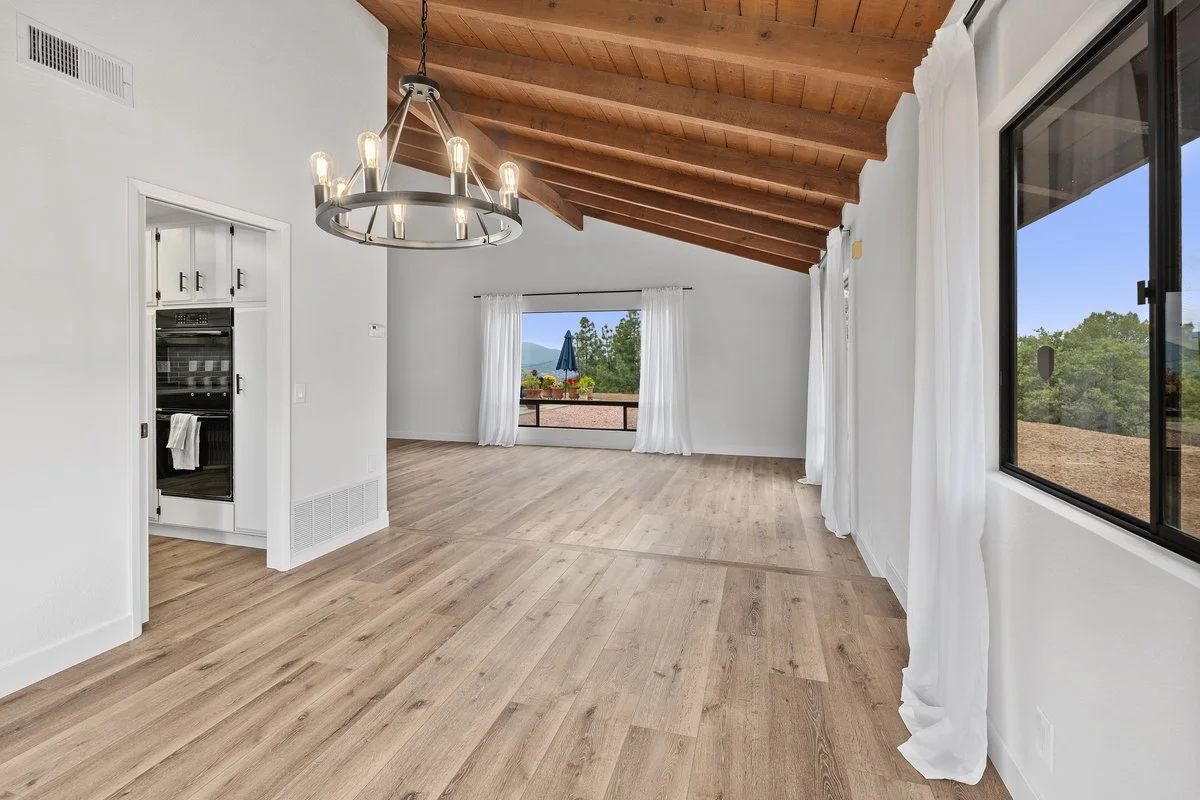 Empty living room with wooden vaulted ceiling, large windows with white curtains, hardwood floors, and a black chandelier