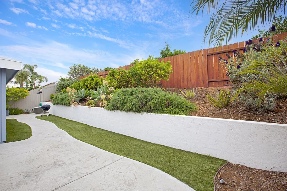 A backyard with a concrete pathway, a grassy strip, a white retaining wall, various green plants and shrubs, a barbecue grill, and a wooden fence under a blue sky with few clouds.