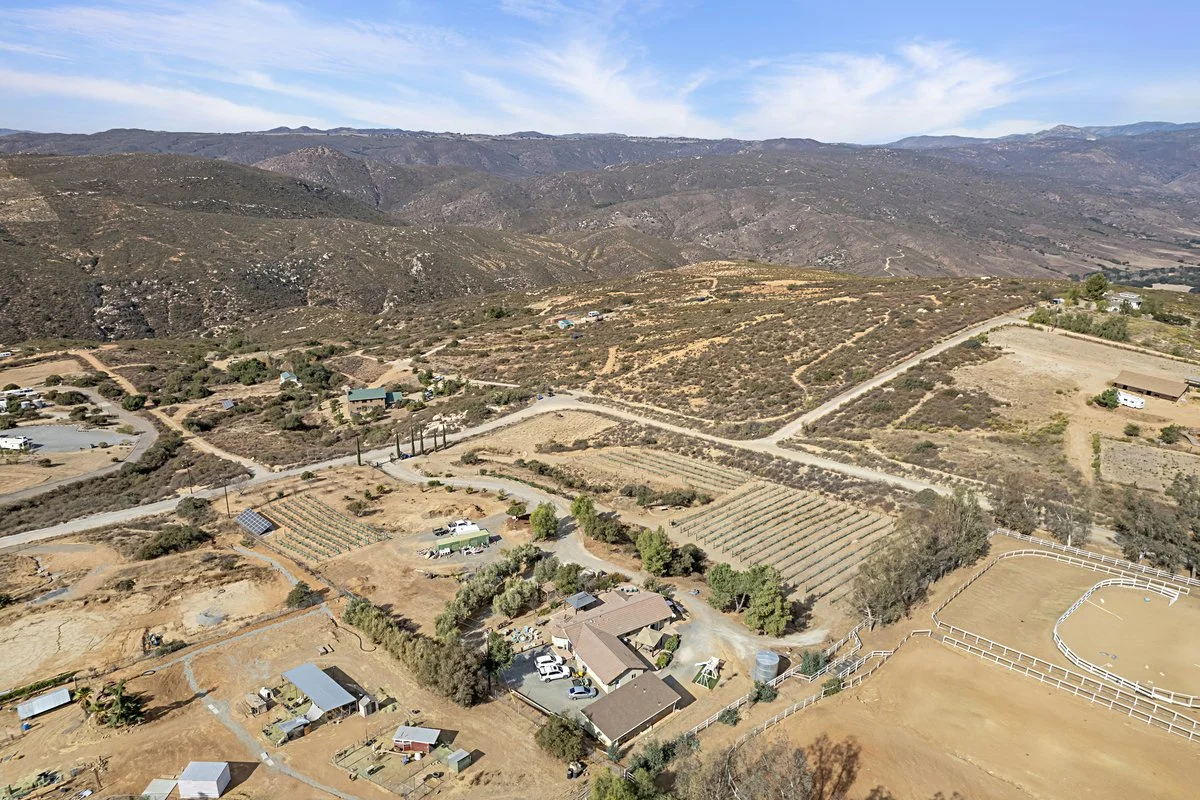 Aerial view of a rural landscape with houses, roads, fields, a small pond, and hilly terrain in the background.