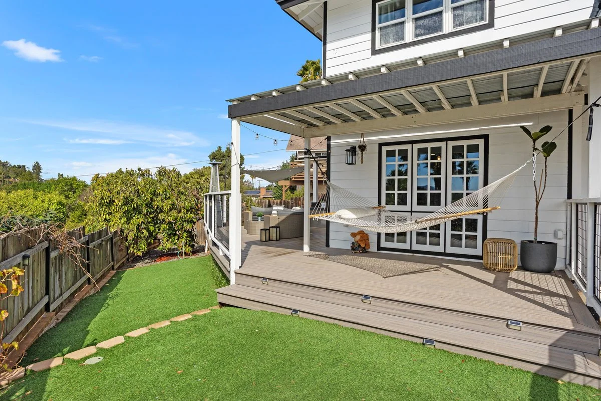 Backyard patio with a hammock, potted plants, and outdoor seating, attached to a white house with black window trim, in sunny weather.