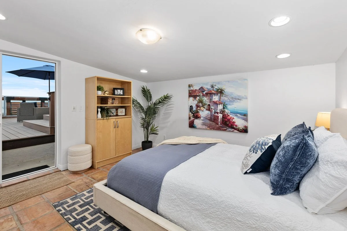 A bedroom with a white bed, dark blue pillows, a painting of a coastal house, a wooden shelf with decor, and a sliding door leading to an outdoor balcony with seating and an umbrella.