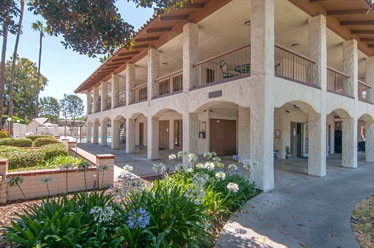 A two-story apartment building with a stucco exterior, wooden roof, balcony on the upper floor, and a pathway with flower beds in front.