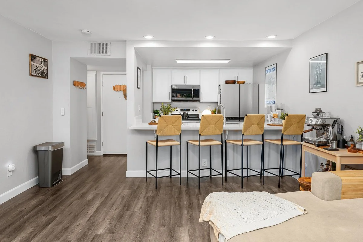 Modern kitchen in an open living space with a breakfast bar and four yellow chairs, white cabinets, stainless steel appliances, and wall art, with a living area and wooden floor.
