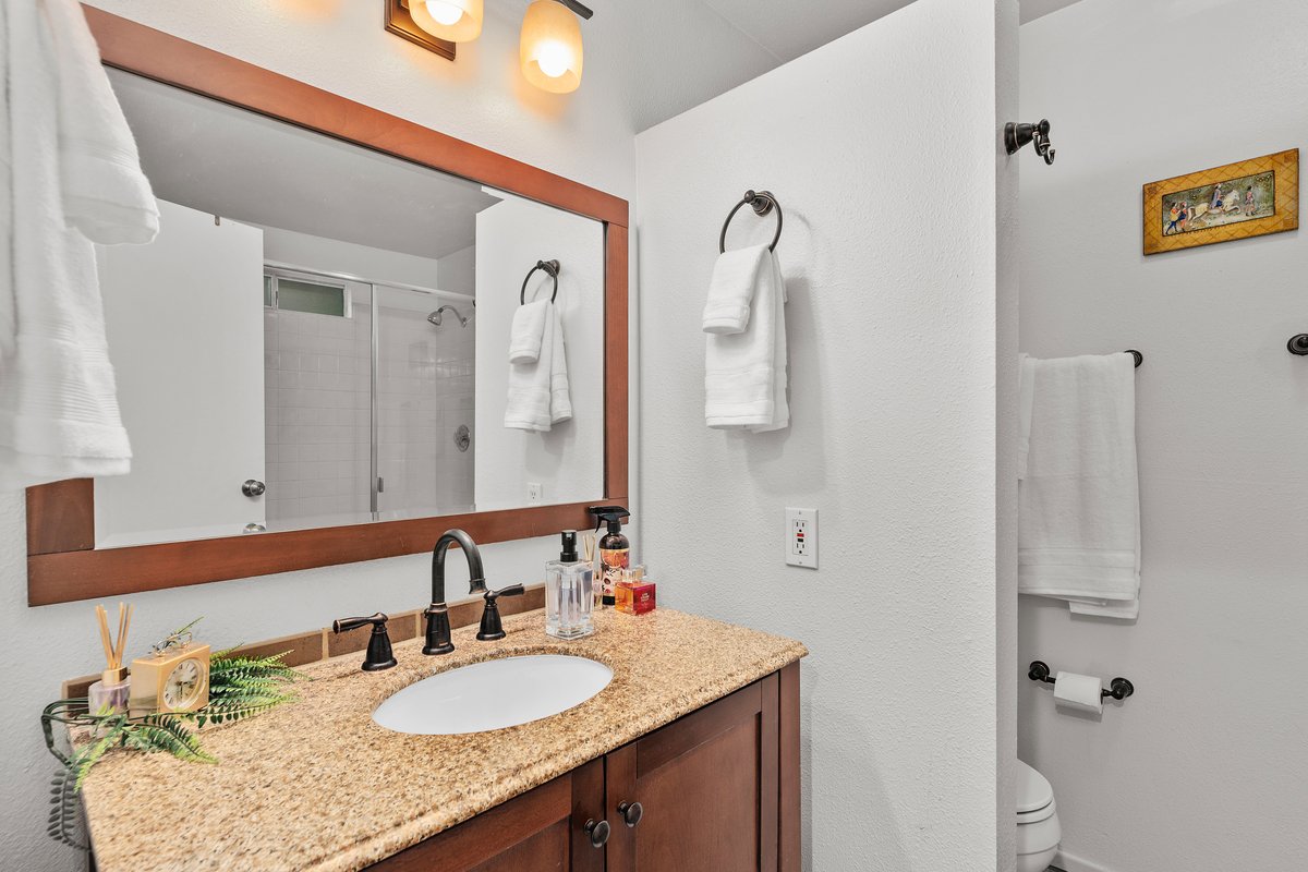 Bathroom vanity with a granite countertop, a large mirror, and two black faucet fixtures. The mirror reflects a shower with a glass door. The vanity area has soap, lotion, and perfume bottles. Towels are hanging on hooks and a towel rack. The bathroo