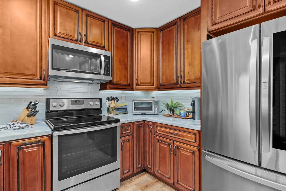 Kitchen with brown cabinets, stainless steel refrigerator, microwave, oven, toaster oven, potted plant, and countertop appliances.