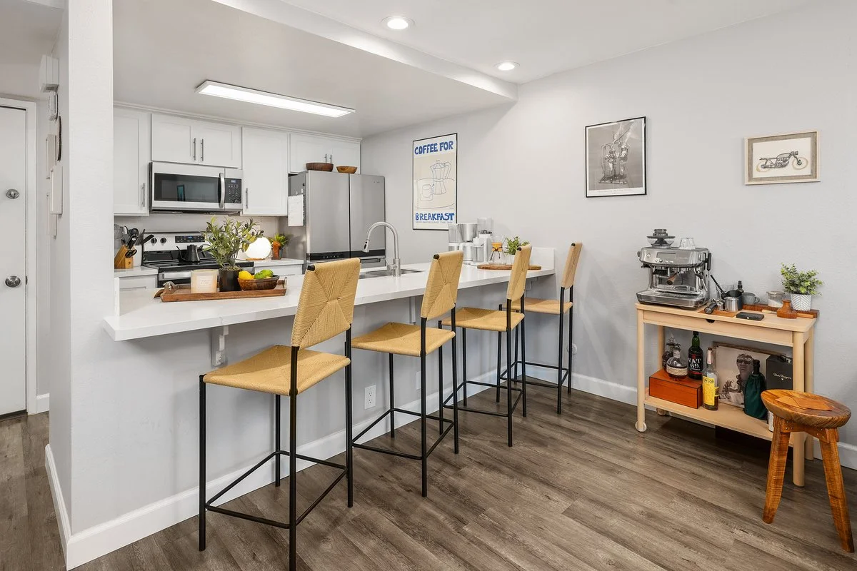 Modern kitchen with white cabinets, stainless steel appliances, and a breakfast bar with four beige high chairs. There are various kitchen decorations and a coffee station on a small wooden side table on the right. Wall art decorates the white walls,