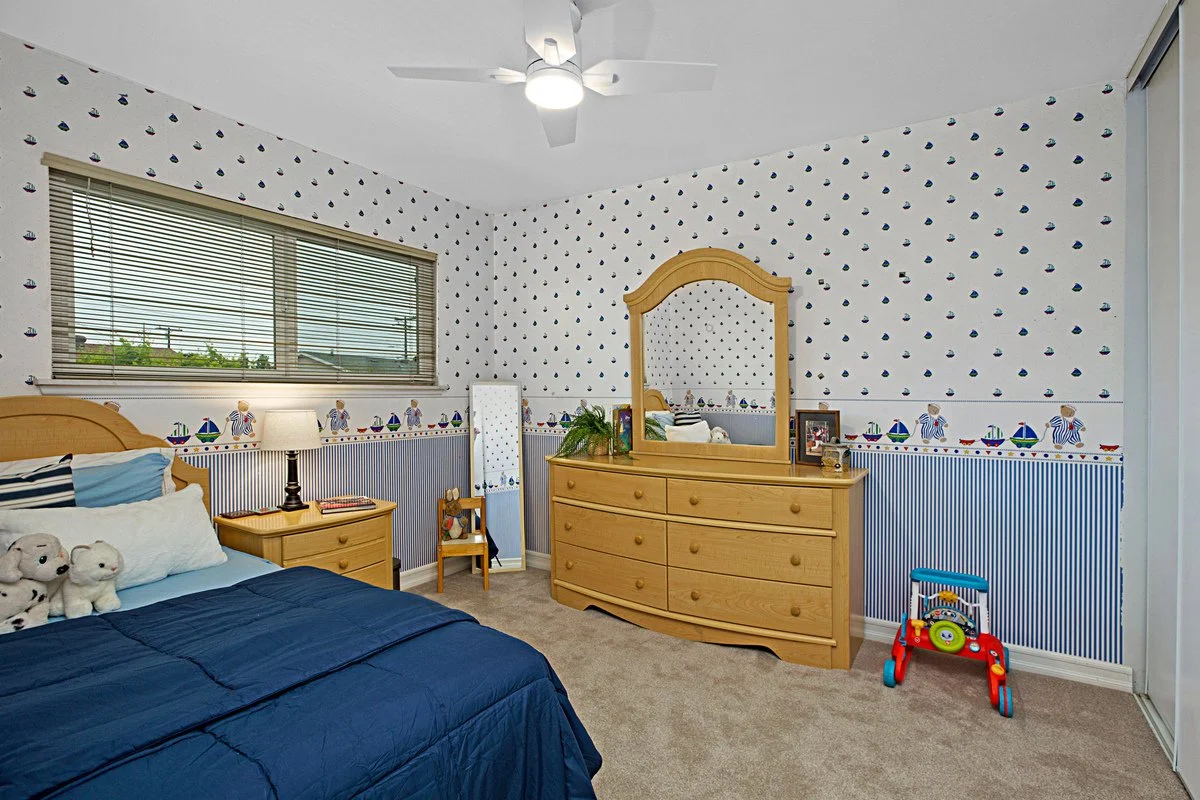 A child's bedroom with nautical-themed wallpaper, a window with closed blinds, a wooden bed with stuffed animals, a nightstand with a lamp, a dresser with a mirror, a small chair, and a red and blue toy wagon.