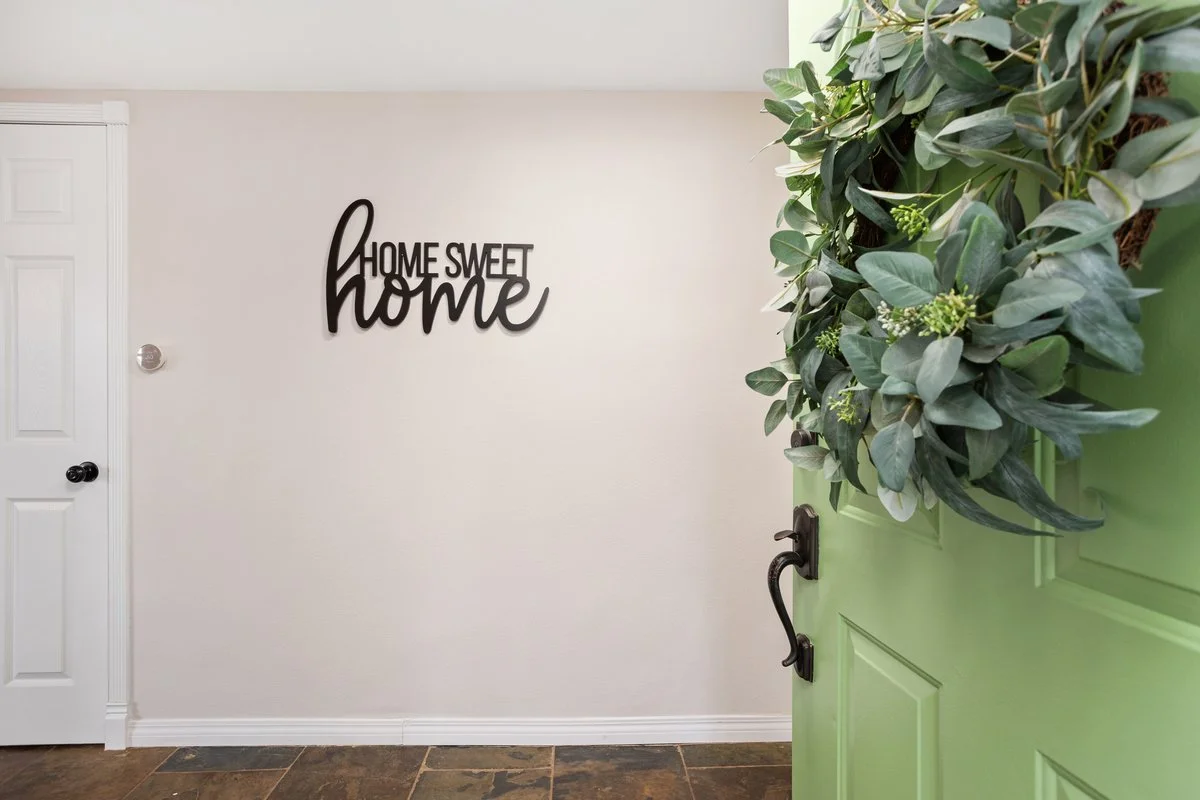 Interior view of a home entrance with a white wall, a decorative sign that says "Home Sweet Home," a closed white door with a black doorknob, and a partially open green door with a black handle and green foliage hanging on it.