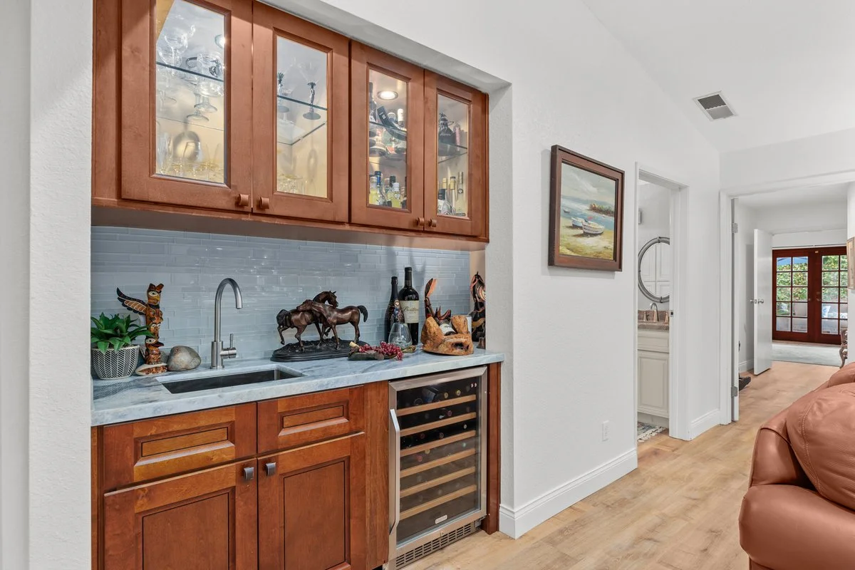 A kitchenette with wooden cabinets, a marble countertop, and a glass-front wine cooler. Decorative items include a plant, horse statues, wine bottles, and a carved wooden sculpture. Adjacent to the kitchenette is a hallway with a framed painting and 