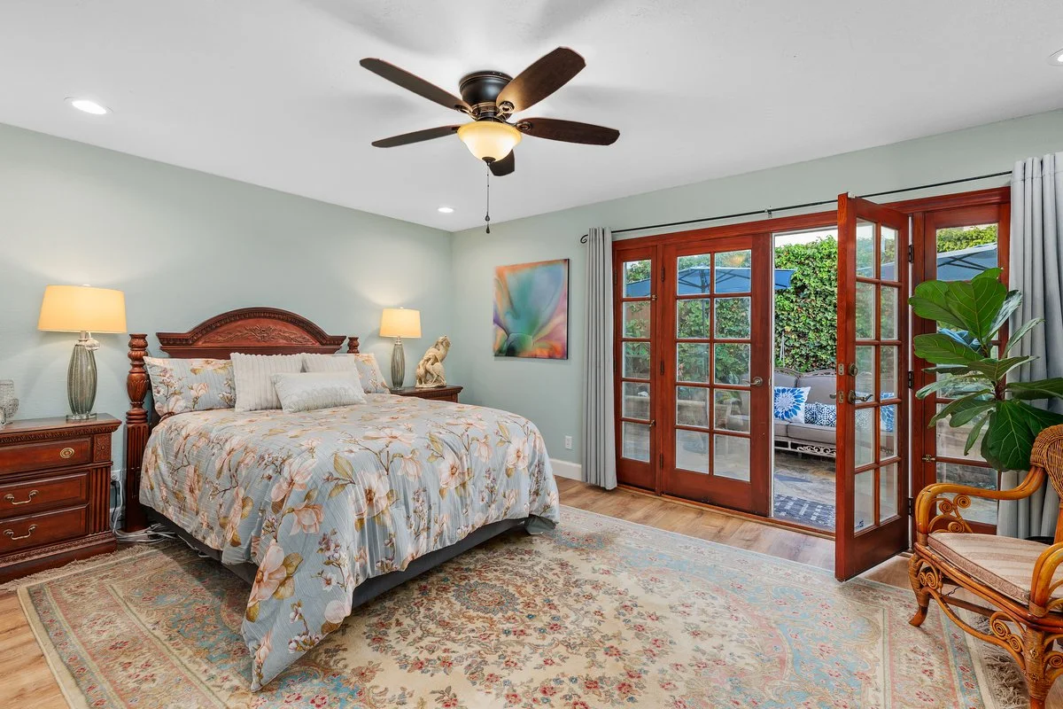A bedroom with a wooden bed, floral bedding, two bedside lamps, a patterned rug, a wooden side table, a painting on the wall, and large glass double doors leading outside to a patio with outdoor furniture and greenery.