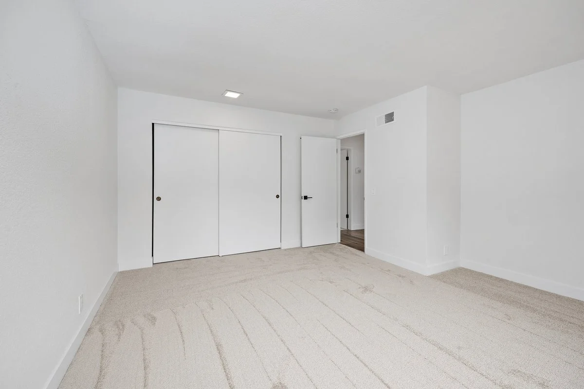 Empty bedroom with white walls, beige carpet, a closet with white sliding doors, and an open door leading to a hallway.