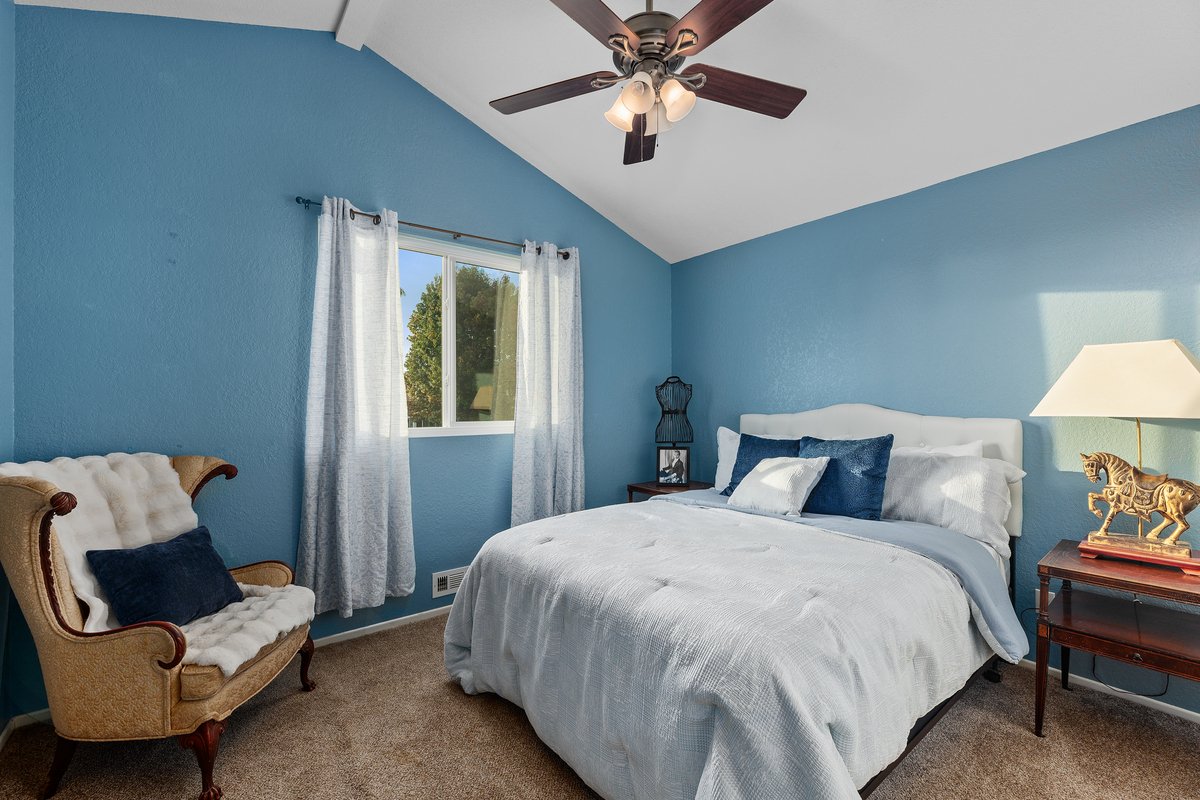 A cozy bedroom with blue walls, a white bed with navy and white pillows, a wooden nightstand with a lamp and decorative horse sculpture, an antique-style chair with a navy pillow, a window with white curtains, and a ceiling fan with lights.