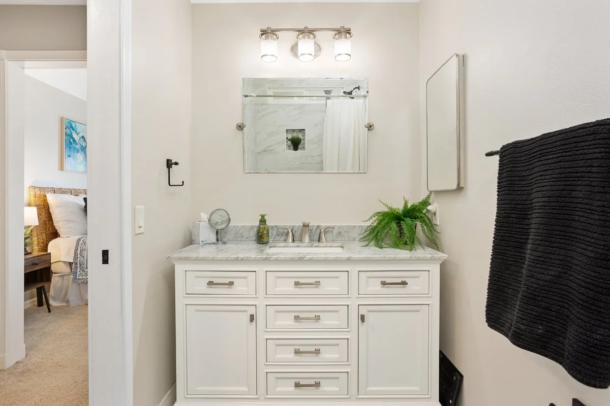 Bathroom with white vanity, marble countertop, mirror, wall-mounted light fixture, green potted plant, black towel, and partial view of a bedroom