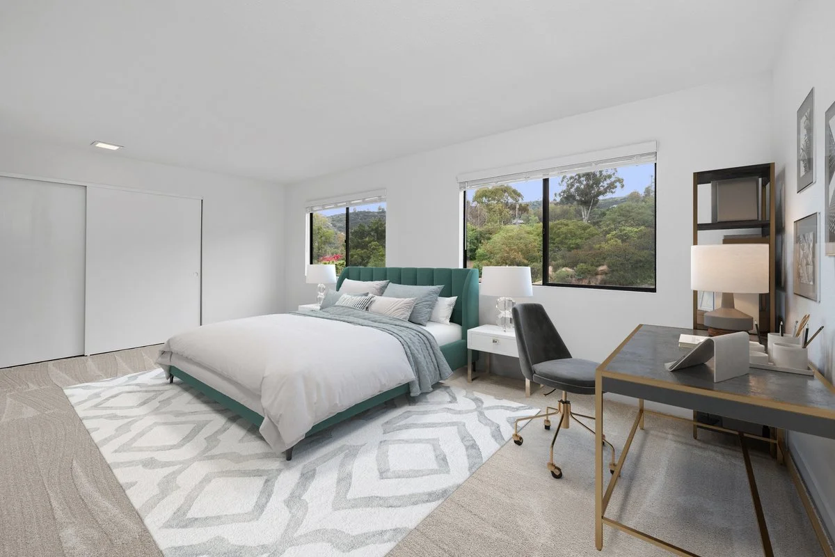 Bedroom with white walls, beige carpet, large bed with teal headboard, white bedding, multiple pillows, white nightstand with lamp, desk with black chair and table lamp, artwork on wall, windows showing greenery outside.