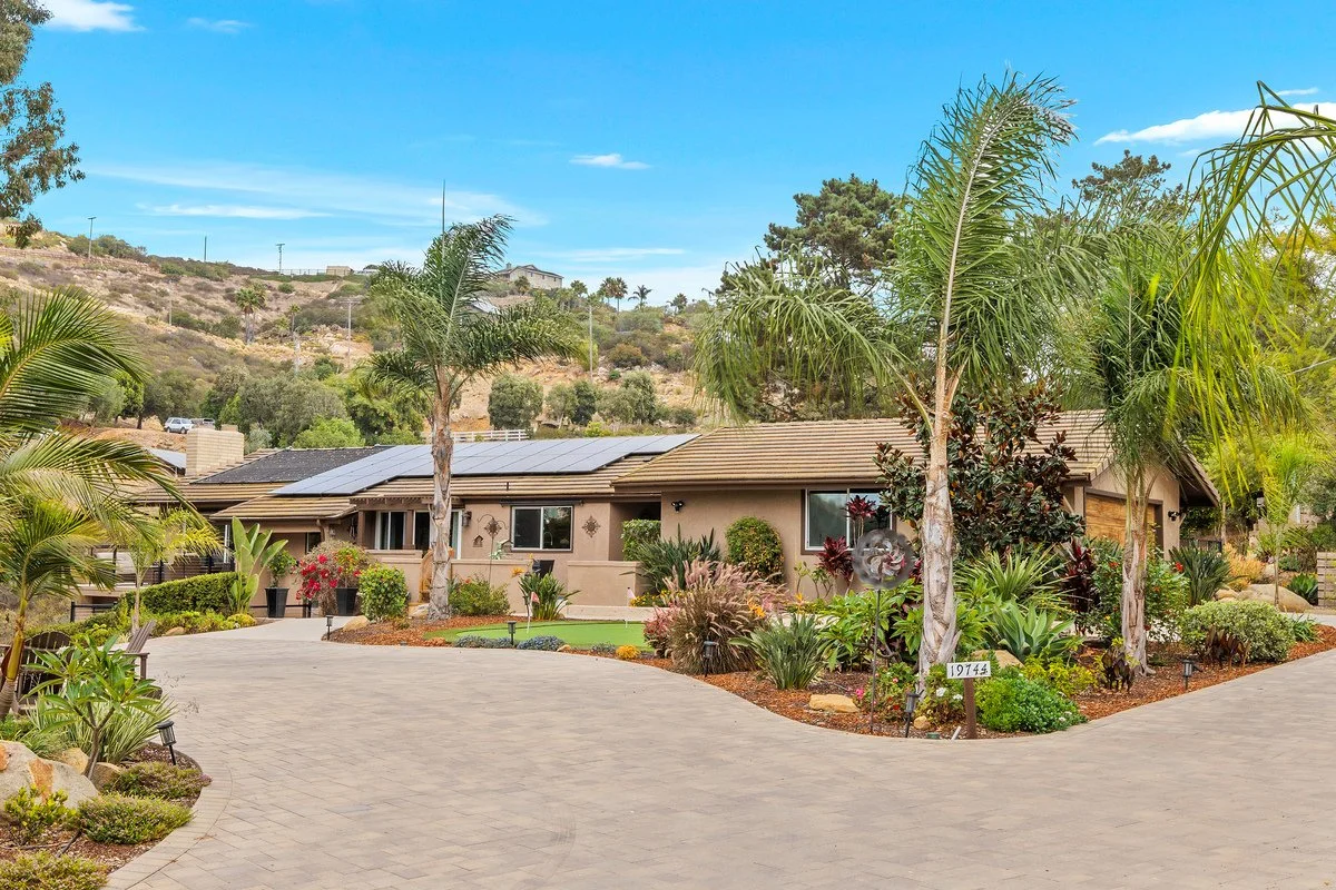 Single-story house with solar panels on the roof, surrounded by desert-style landscaping, palm trees, and a curved driveway, on a sunny day with blue sky.
