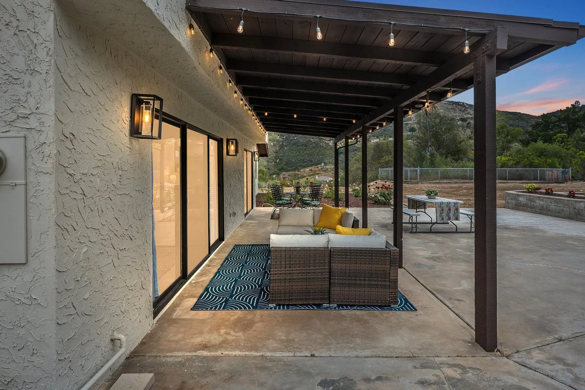 Outdoor patio at sunset with a section of white stucco house, string lights, a wicker couch with yellow pillows on a blue patterned rug, a table, and distant mountains.