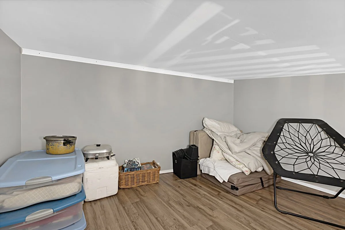 A cluttered room with a gray wall, a bed with disorganized blankets and pillows, a spider web-shaped hanging chair, stacked plastic storage bins, a laundry basket, a small grill, a wicker basket, and a few black storage containers on a hardwood floor