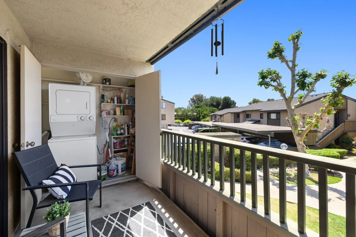Apartment balcony with laundry area, black chair with striped pillow, small table with potted plant, wind chime hanging, overlooking a courtyard with trees, cars, and neighboring buildings on a sunny day.