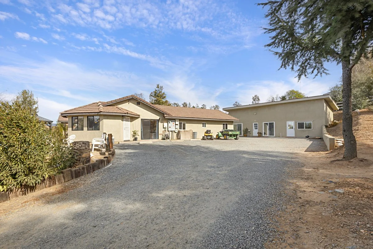 A large gravel backyard with a house on the left and a building on the right, trees on the edges, and a blue sky with some clouds overhead.