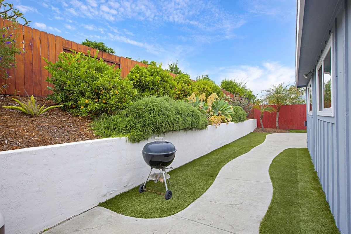 Backyard with a winding concrete pathway, green grass borders, a white retaining wall with lush bushes and plants, a black charcoal grill, and a blue house with white trim. A red wooden fence surrounds the yard under a partly cloudy sky.