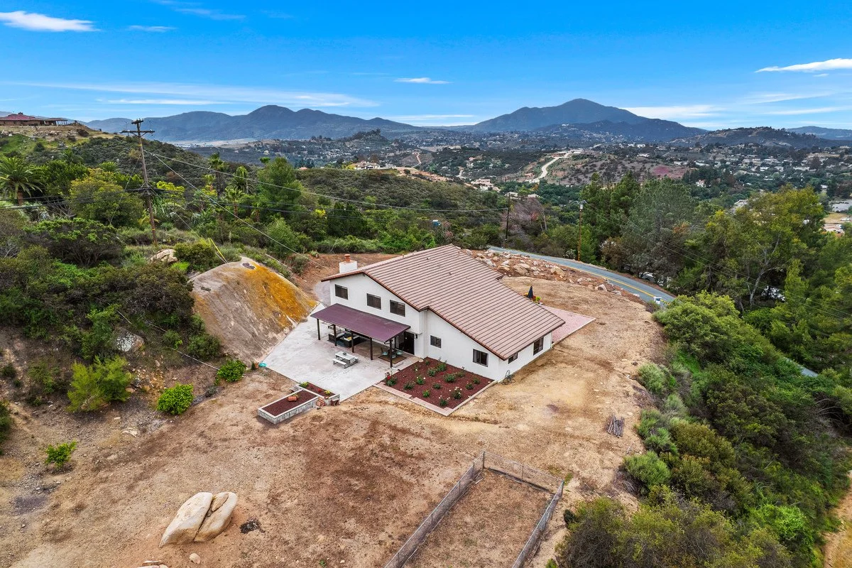 A house sits on a hill with a mountain range in the background, surrounded by trees and dry terrain, with a partly cloudy sky overhead.