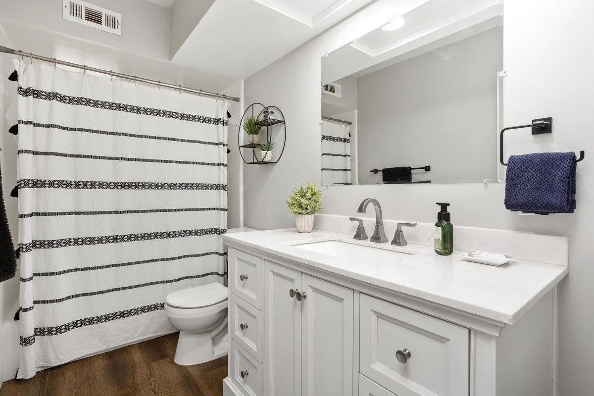 A modern bathroom with white cabinets and a marble countertop. A large mirror reflects part of the room. There is a shower with a black and white striped curtain, a toilet, and a wall-mounted black towel rack holding a navy towel. A small round shelf