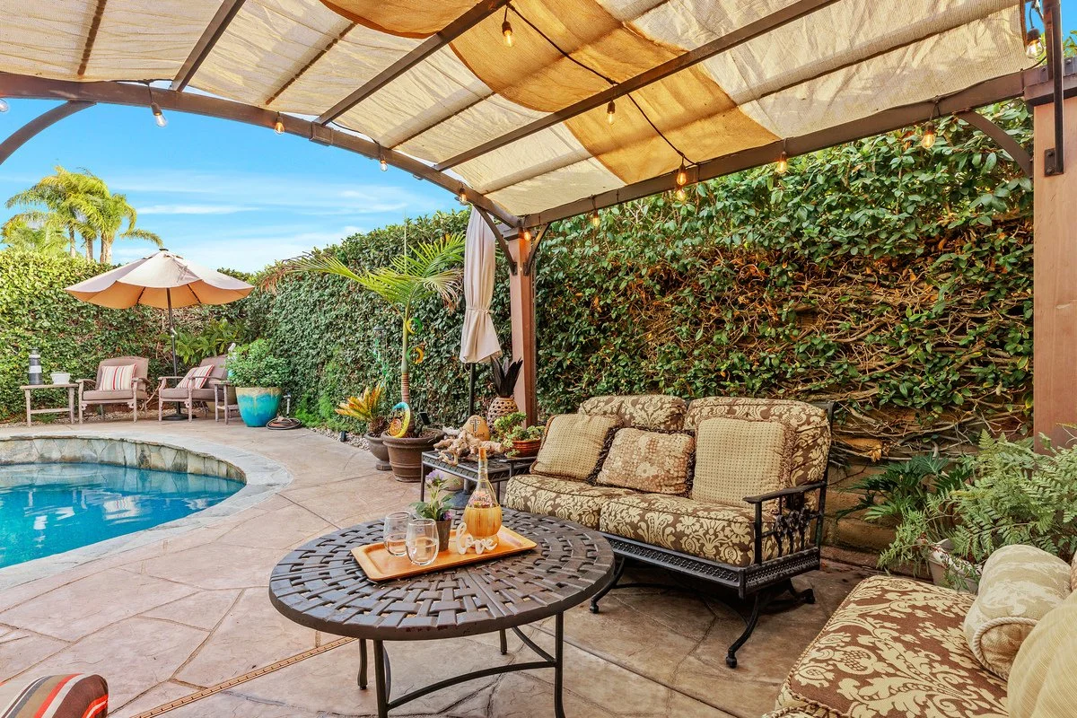 Outdoor poolside patio area under a canopy with seating, potted plants, and string lights, overlooking a lush green hedge and blue sky.