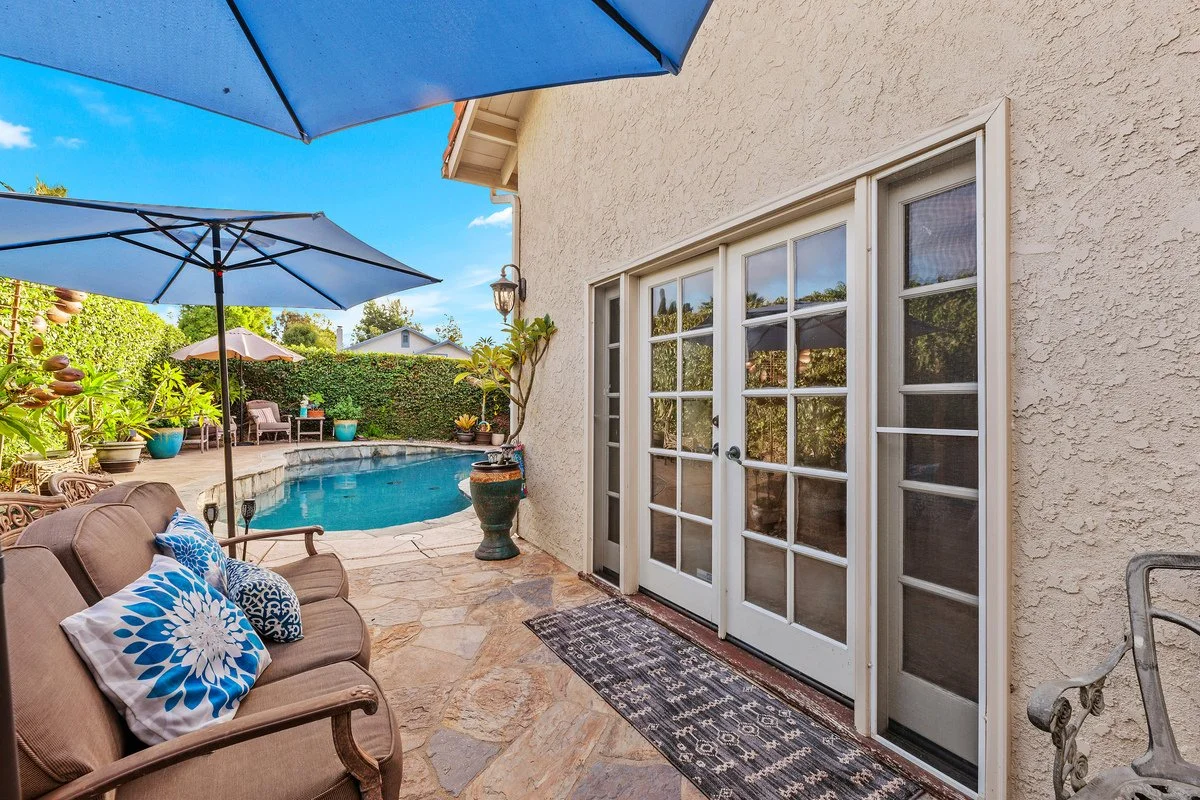 Backyard patio with a swimming pool, two blue umbrellas, and outdoor seating including a cushioned sofa with patterned pillows, potted plants, and a sliding glass door.