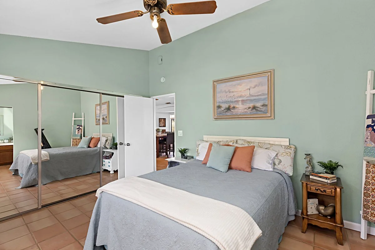 A bedroom with a queen-sized bed with multiple pillows, a nightstand with decorative items, a wall painting above the bed, a ceiling fan, and a large mirrored closet door.