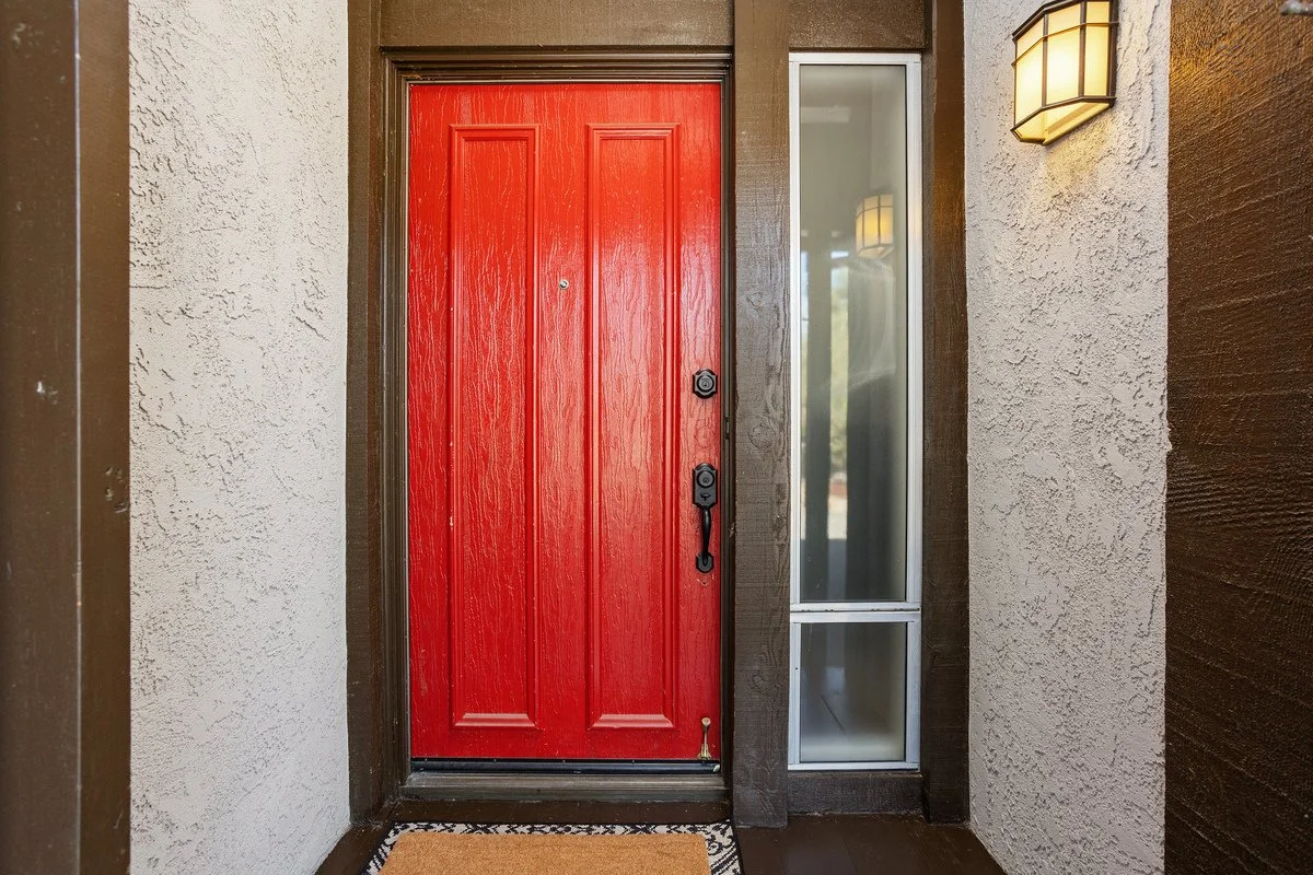 Red front door with black handle and deadbolt, next to a glass side panel and a wall-mounted light fixture.