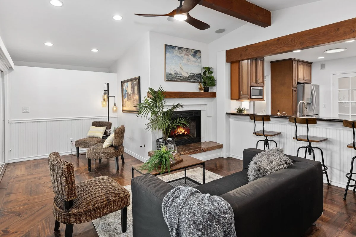 Living room with white walls, hardwood flooring, a black sofa with a gray blanket and pillow, a small wooden coffee table with plants, a fireplace with artwork above, two wicker chairs, a floor lamp, and a kitchen area with bar stools and wooden cabi