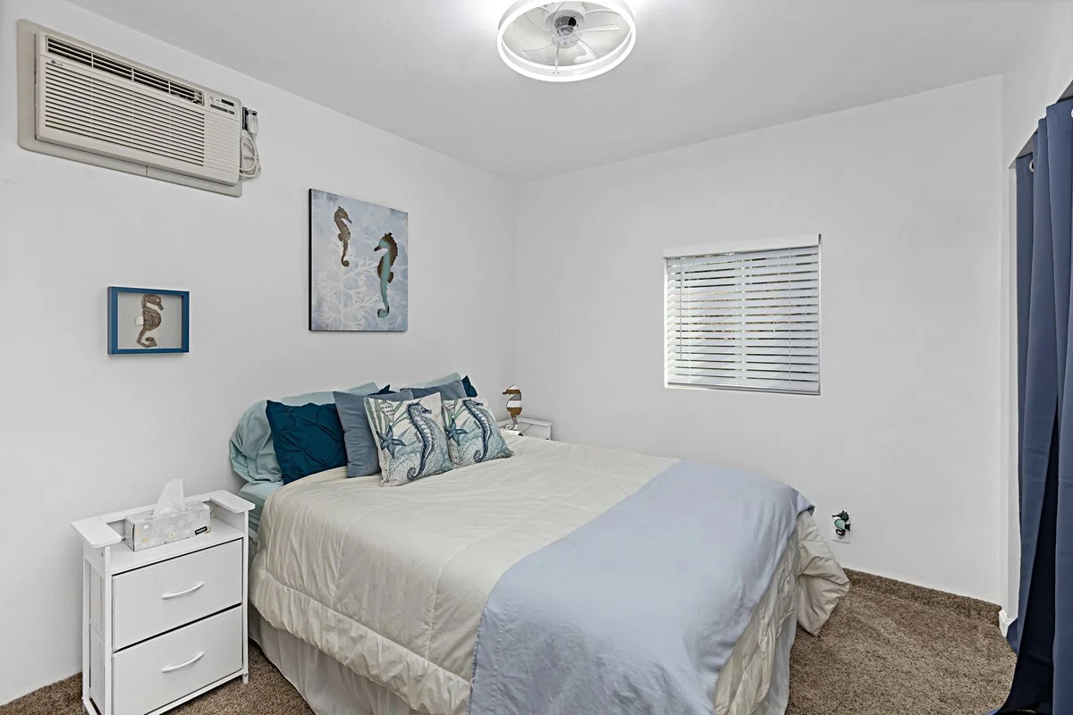 A bedroom with white walls, a white bedspread, pillows with seahorse designs, a small white nightstand with tissues, a window with blinds, and a blue curtain. There is an air conditioning unit above the bed and a ceiling fan.