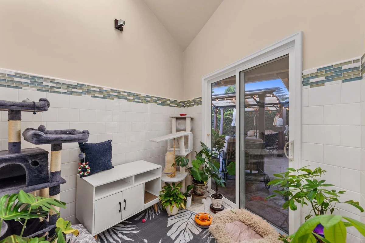 Indoor space with plants, a white cabinet, a dark gray cat tree, a cat scratching post, a furry pet bed, and sliding glass door leading outside patio with outdoor furniture.